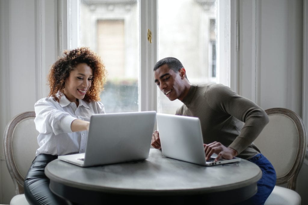 positive young ethnic colleagues using laptop on round table near window at home